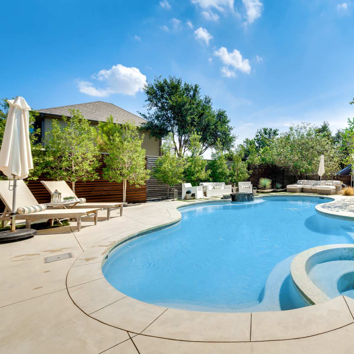 A residential backyard featuring a large, shaped swimming pool with a spa area, surrounded by greenery and patio furniture.