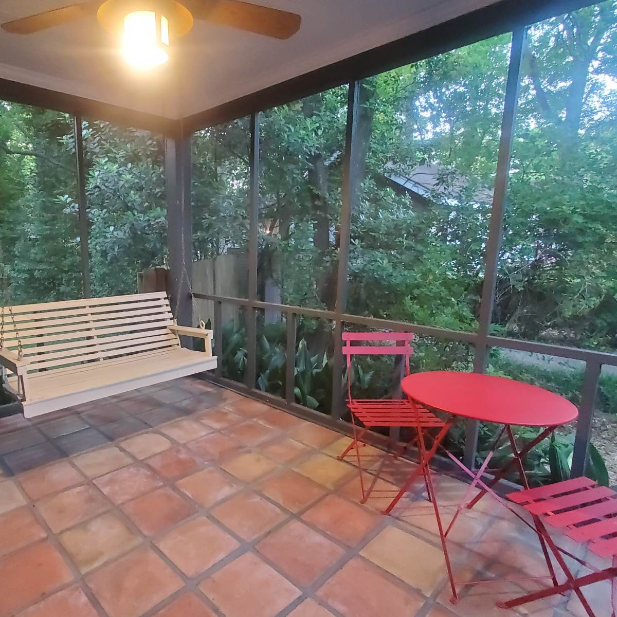 A screened porch with a wooden swing and a small red table with two matching chairs, surrounded by greenery.
