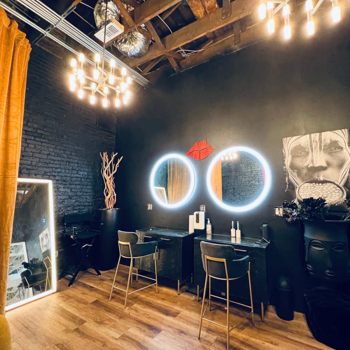 A modern black-themed salon interior featuring two circular mirrors with LED lighting, a dark wall art piece, a decorative plant, and elegant seating.