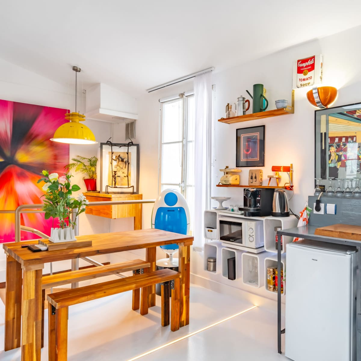 A bright, modern kitchen setup featuring a wooden dining table and chairs, colorful wall art, and a compact kitchen area with a sink and storage shelves.