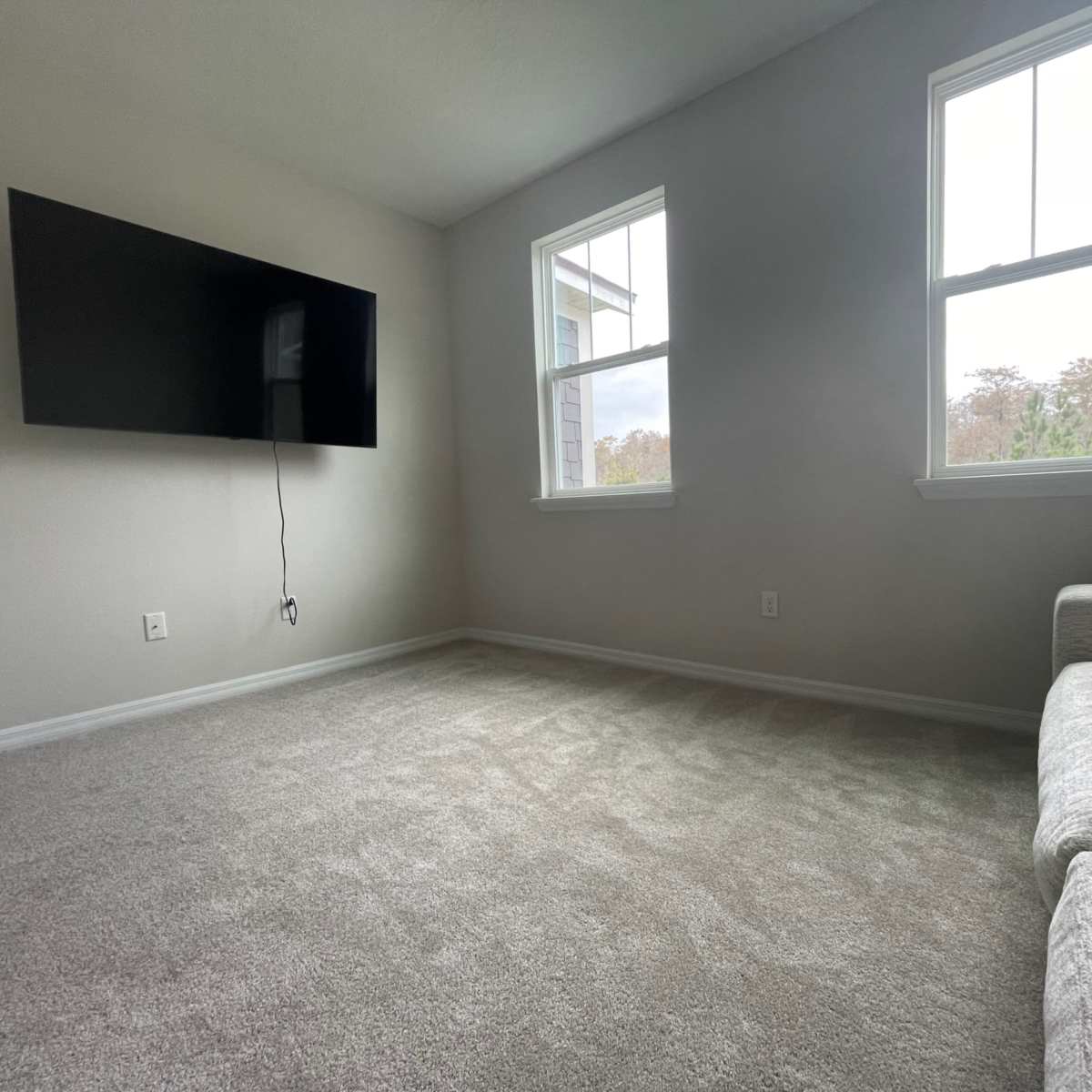A sparsely furnished living room with gray carpet, a wall-mounted television, two windows, and a light-colored sofa.
