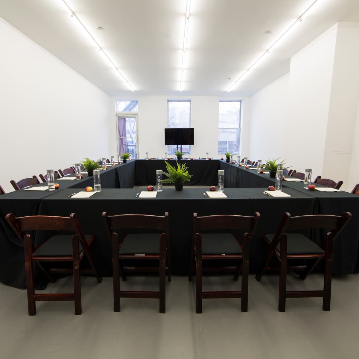 A long table is set up in a spacious, brightly lit room with chairs arranged around it and a television mounted on the wall.