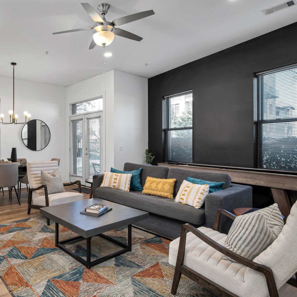 A modern living room with a sofa, chairs, a coffee table, and a dining area, featuring large windows and a geometric-patterned rug.