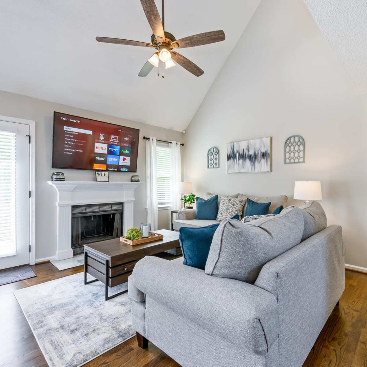 A living room features a gray sectional sofa, a wooden coffee table, and a mounted television above a fireplace, all illuminated by natural light.
