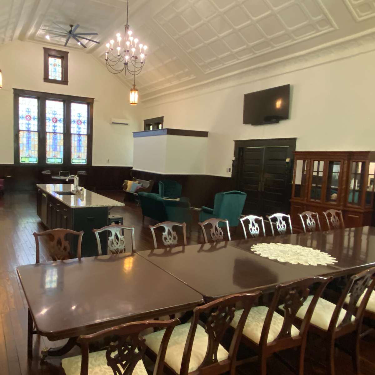 A spacious room with high ceilings, stained glass windows, and a long dining table surrounded by chairs, alongside a kitchen area and seating arrangements.