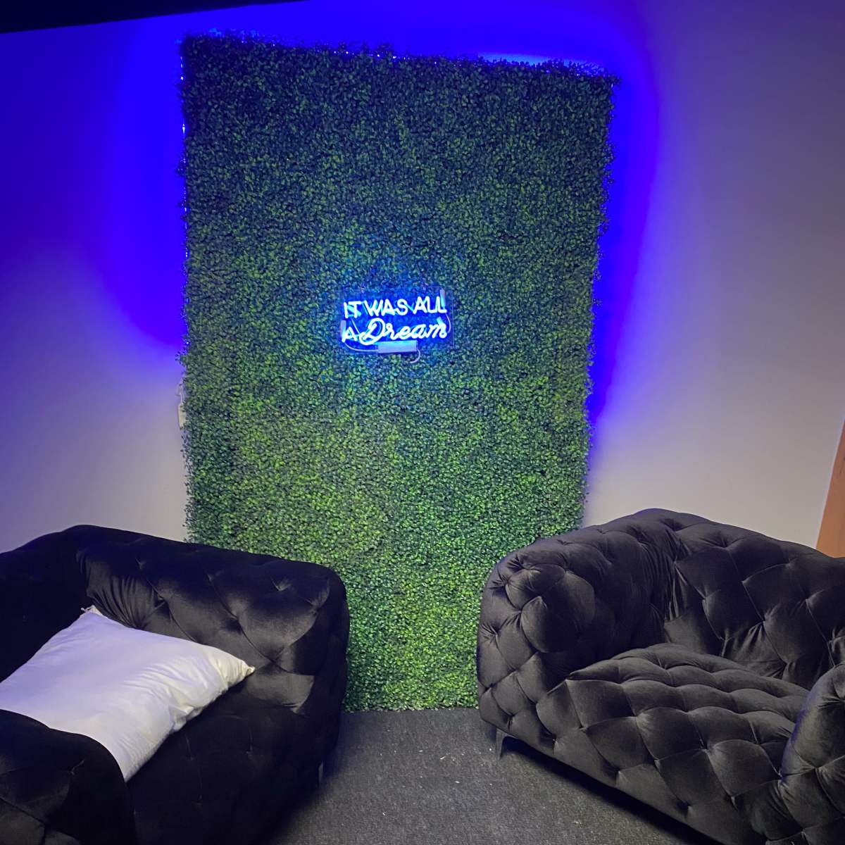 A small indoor space featuring two black tufted sofas facing a vertical green wall with a neon sign that reads, "IF IT WAS ALL A DREAM."