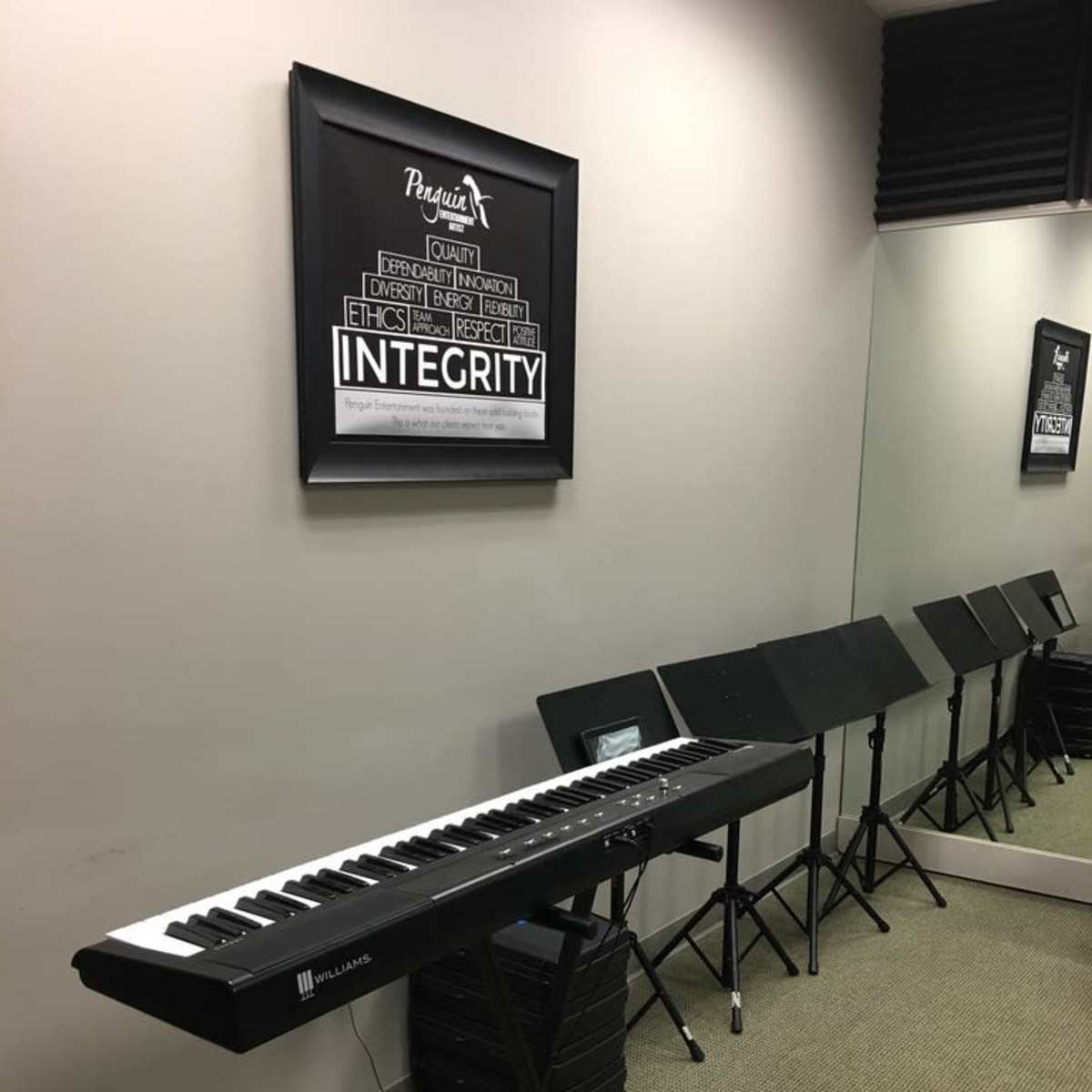 A digital keyboard on a stand in a music practice room, with sheet music stands lined up against the wall and a framed poster displaying the word "INTEGRITY."