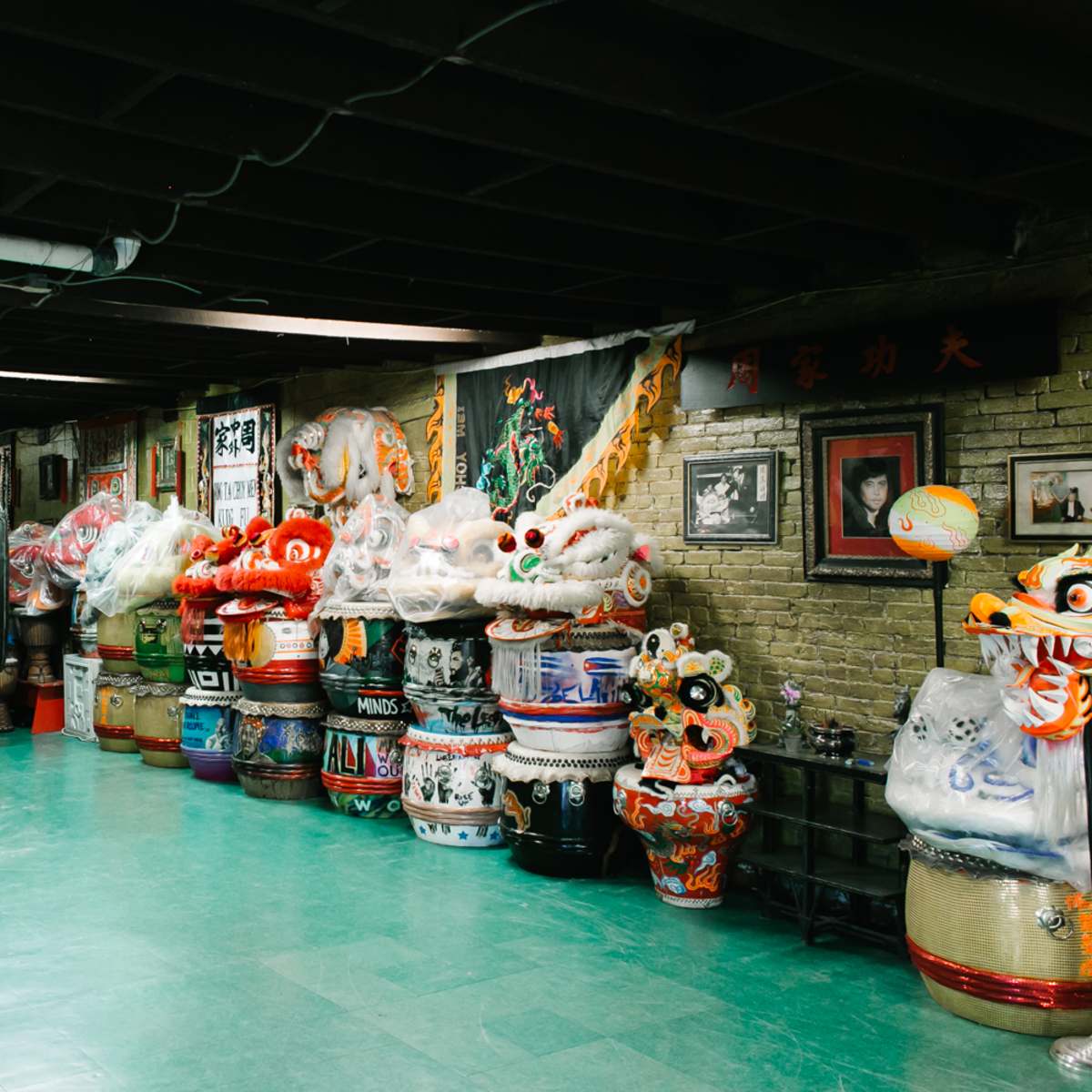 A dimly lit room filled with colorful dragon and lion dance costumes and masks along the walls and stacks of decorative drums.
