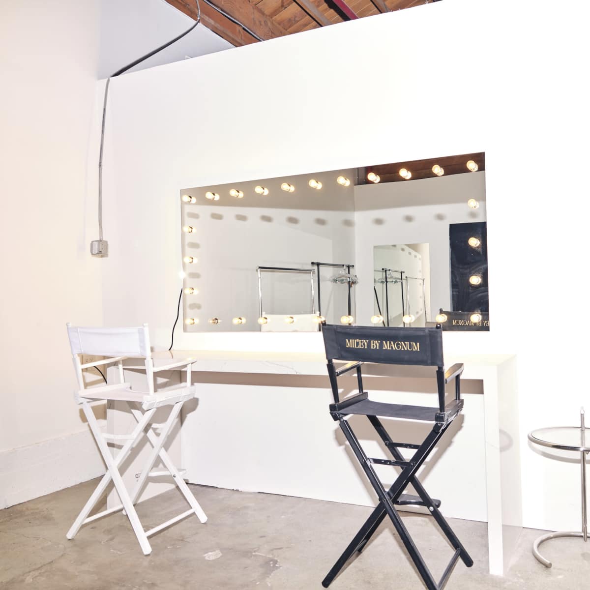 A minimalist makeup room features a large mirror surrounded by lights, two director's chairs, and a small side table.