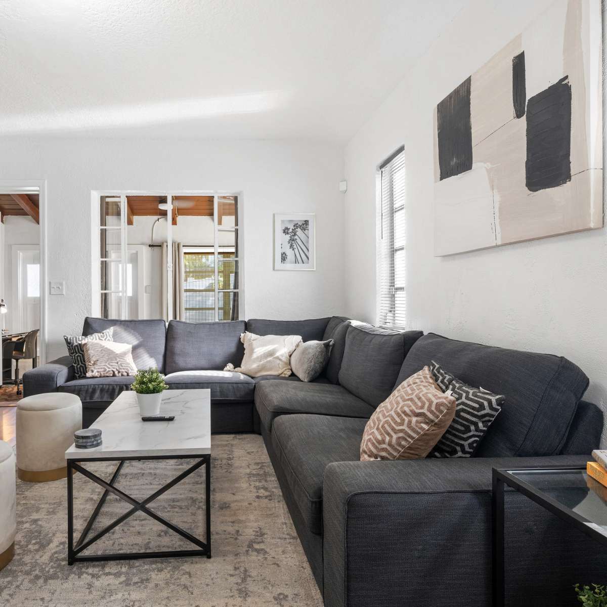 A modern living room features a dark sectional sofa, a glass-topped coffee table, and a combination of decorative stools and plants.