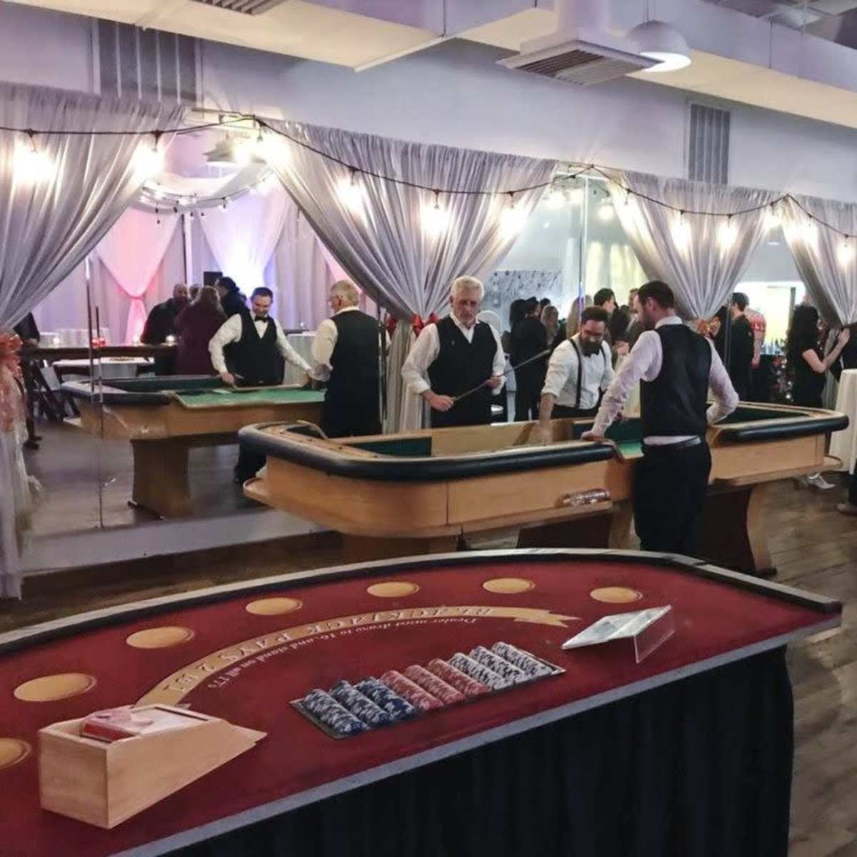 The scene depicts a bustling casino-themed event with guests mingling around gaming tables in a well-decorated room.