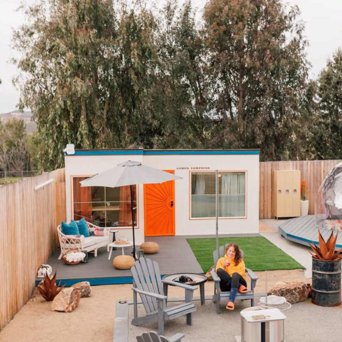 A woman sits outside a colorful modern cabin next to a geodesic dome in a landscaped outdoor area with seating and greenery.