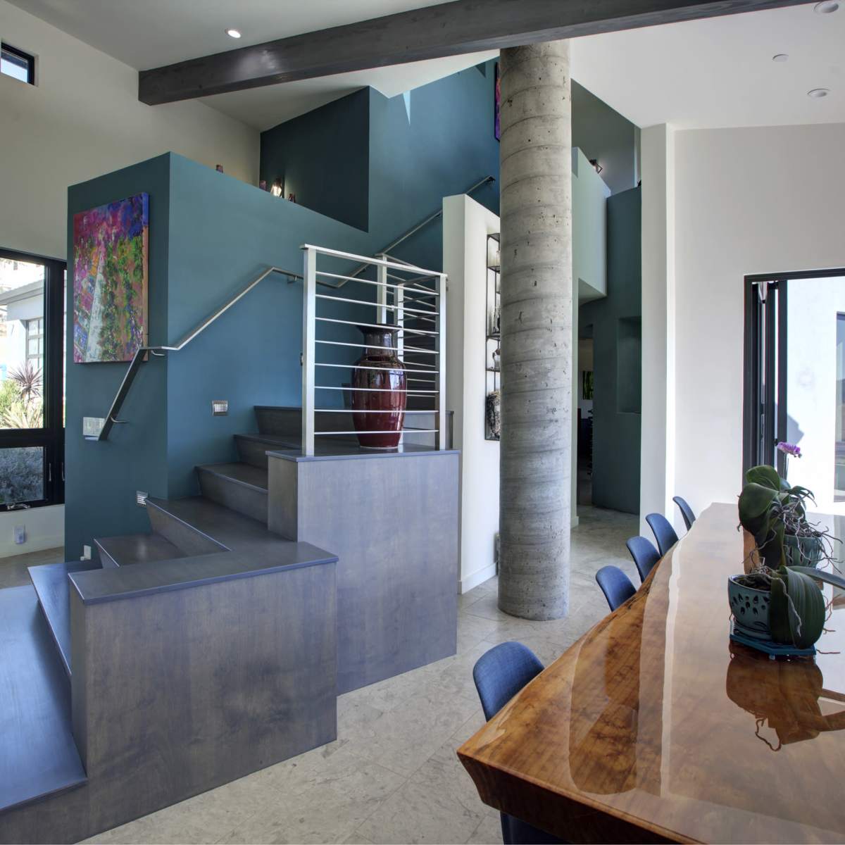 A modern interior space featuring a staircase with a railing next to a dining table, framed by large windows that allow natural light to enter.