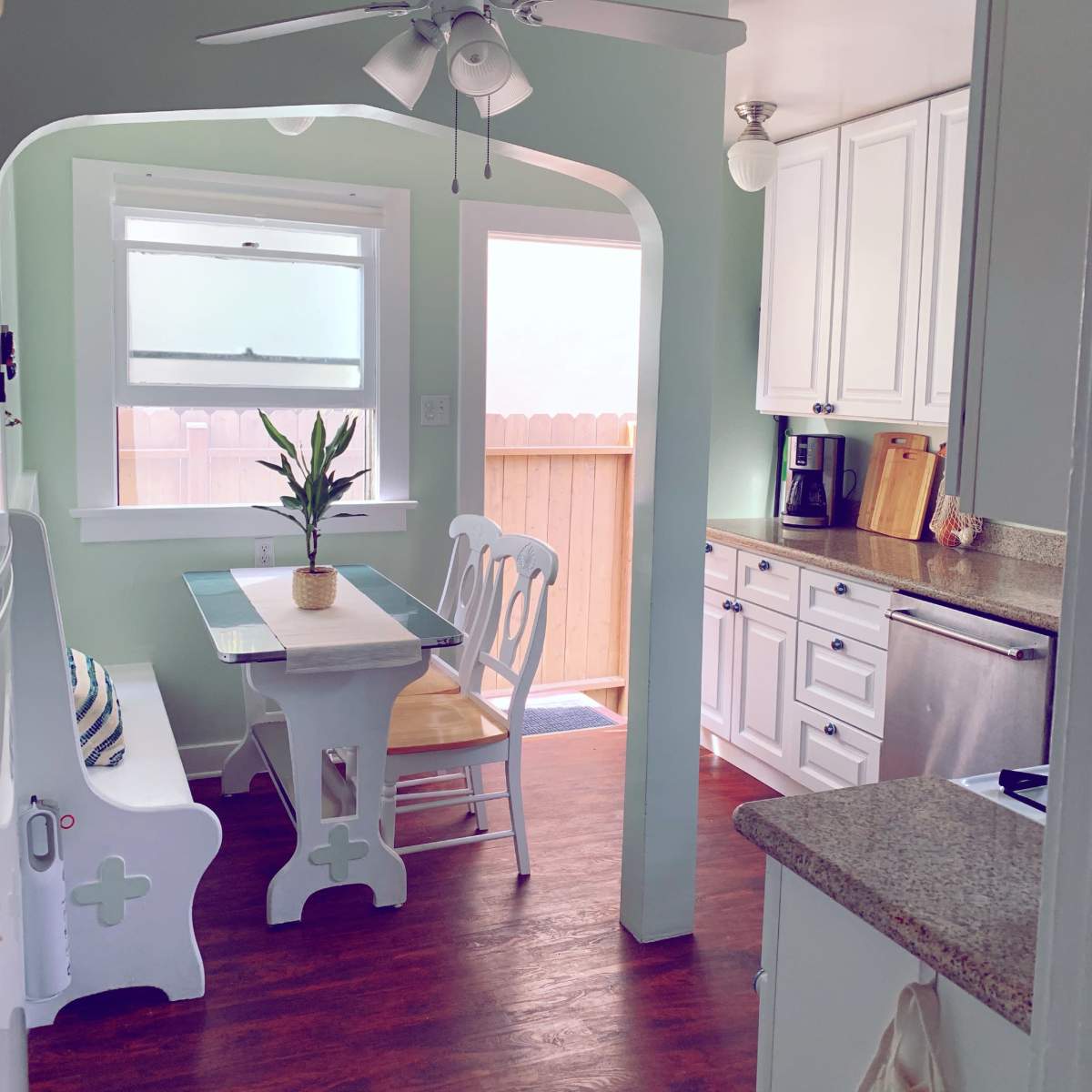 A small kitchen area featuring a white table with a potted plant, a bench, and white cabinets, with natural light coming through a window.