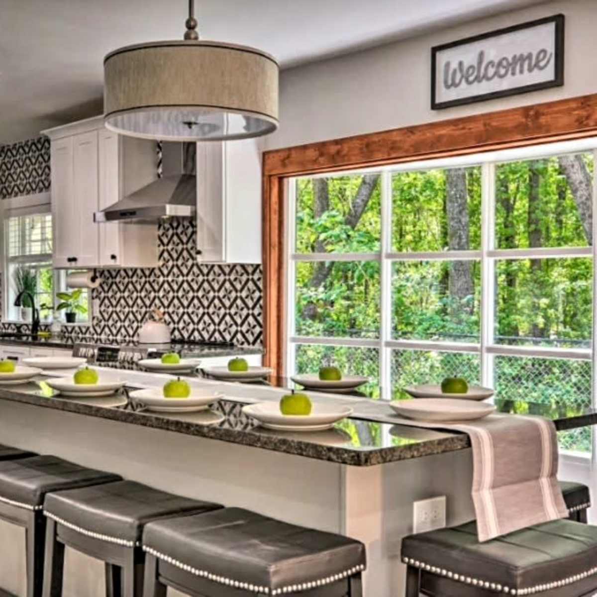 The kitchen features a modern design with white cabinetry, a large island topped with green apples, and a window overlooking a wooded area.