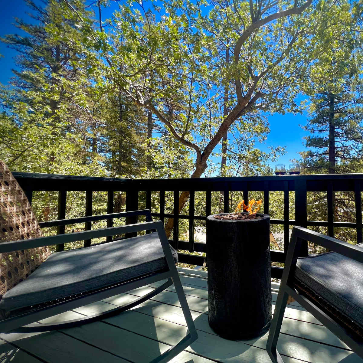 A deck with two black chairs and a small fire pit is surrounded by trees under a clear blue sky.