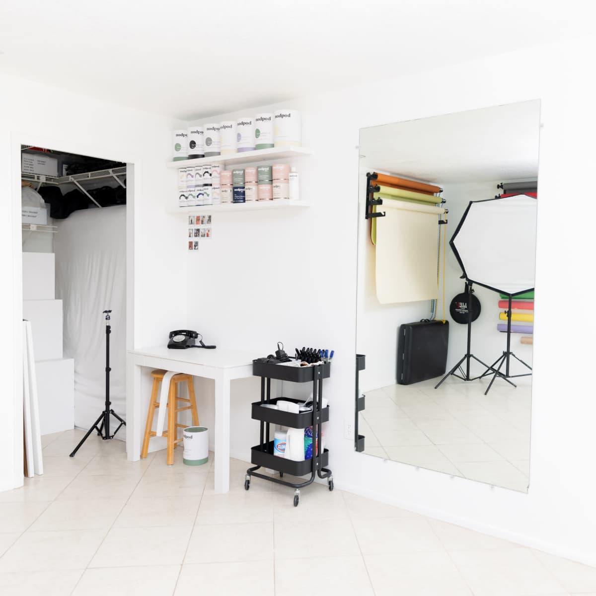 A bright, minimalistic room with a large mirror, a small cart with art supplies, a telephone, and photography equipment in the background.