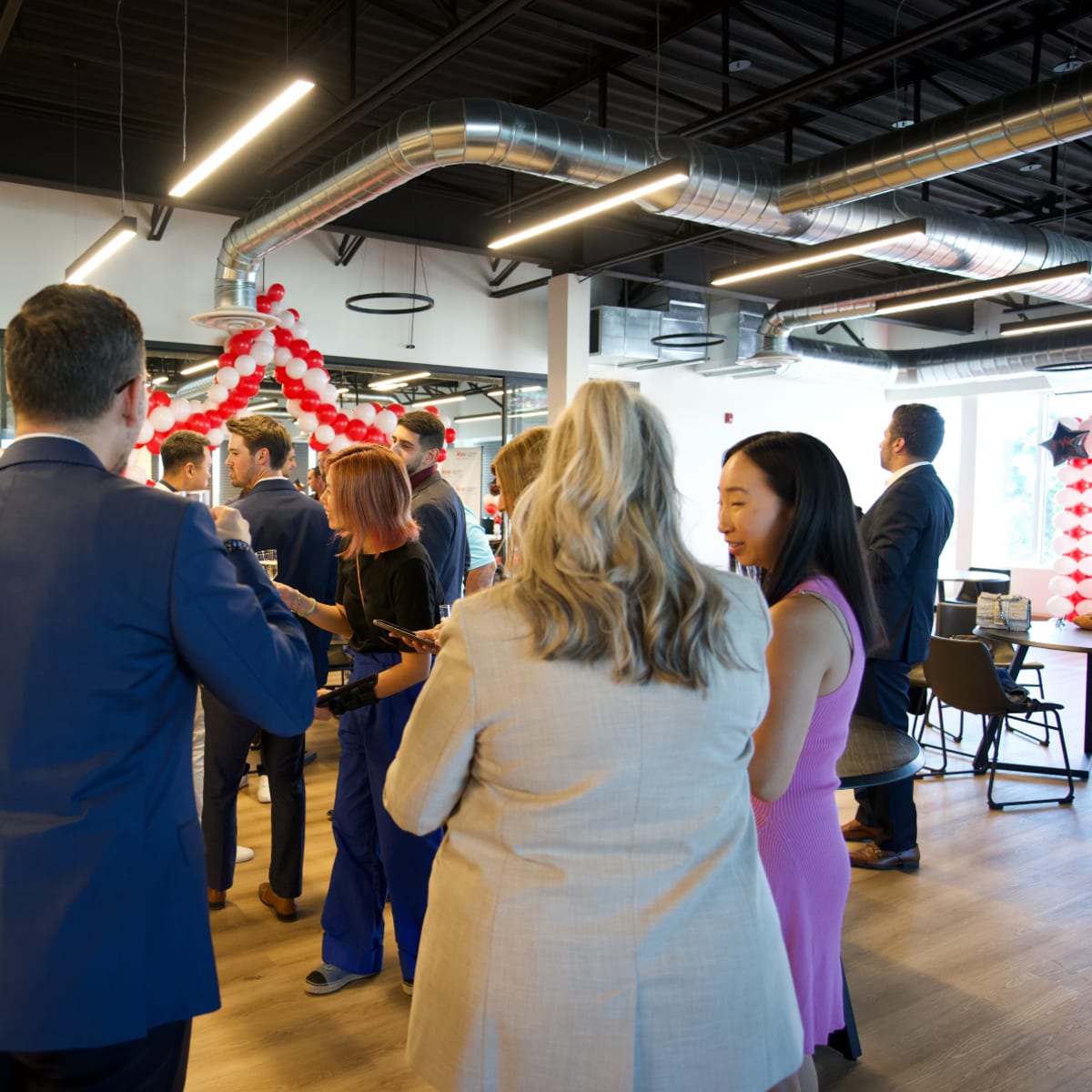 A group of professionals gathers around a buffet table in a modern office space decorated with red and white balloons.