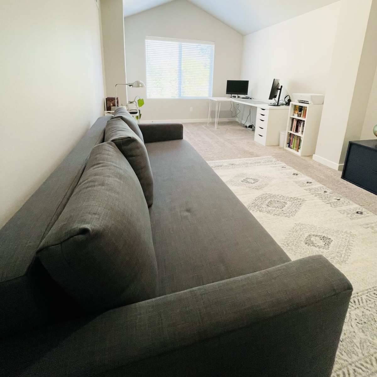 A gray sofa is positioned in a room with a peaked ceiling, a window providing natural light, and a work desk with a computer.