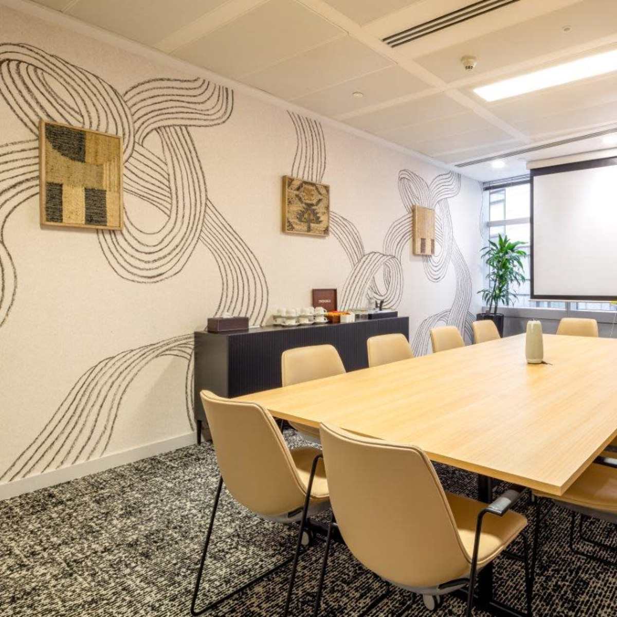 A conference room is set with a large wooden table surrounded by beige chairs, featuring a projector screen and decorative wall art.