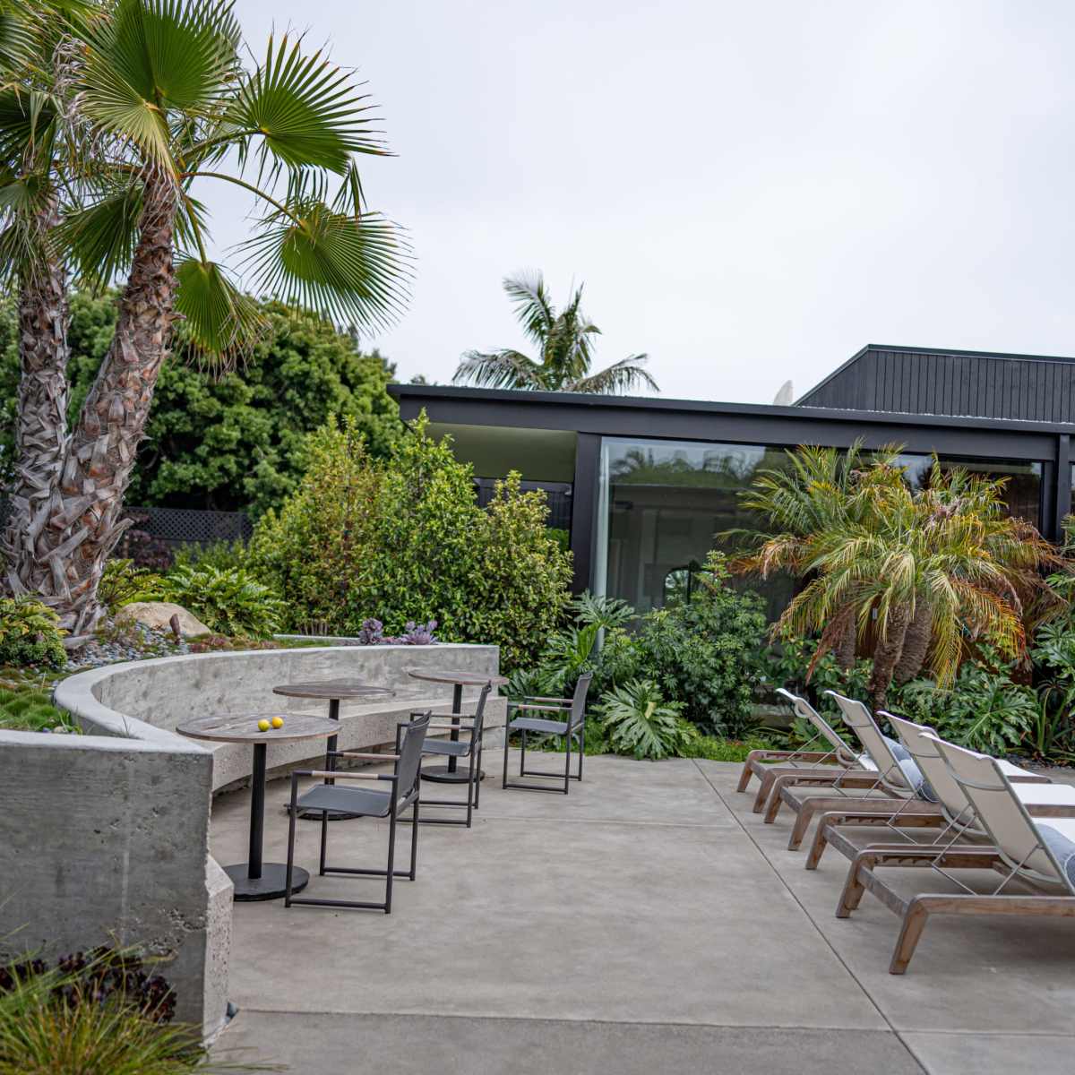 A modern outdoor area features a poolside seating arrangement with loungers and a concrete table set amidst lush tropical landscaping.