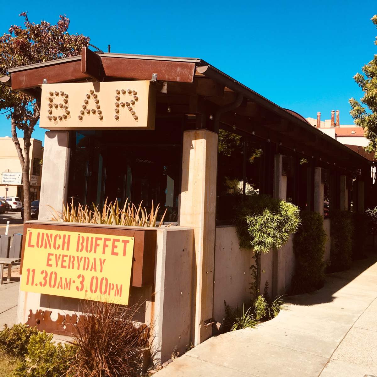 A building with a large sign reading "BAR" and a poster advertising a lunch buffet is located on a clear day with a blue sky.