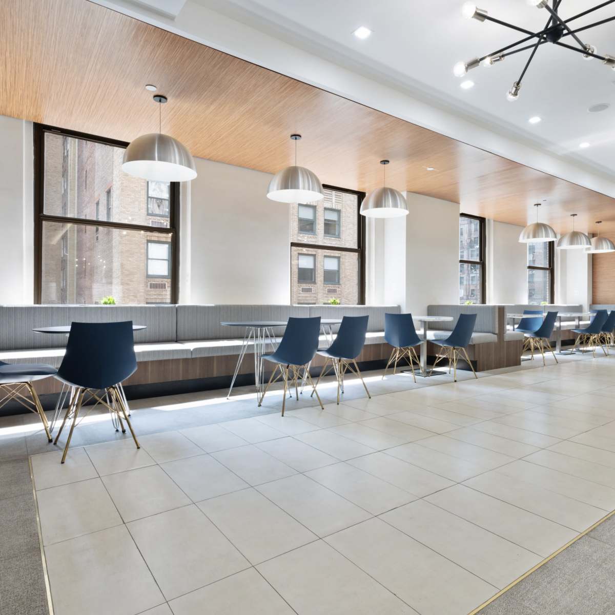 A modern indoor dining area featuring long communal tables with blue chairs, large windows, and pendant lighting.