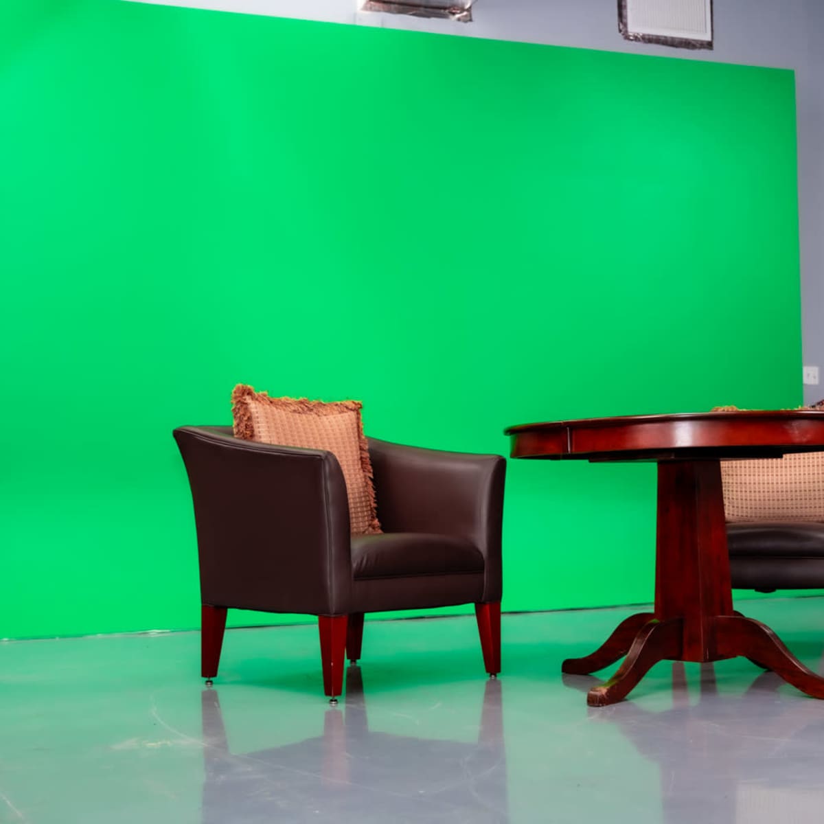 Two dark leather chairs with decorative cushions positioned around a wooden table in front of a green screen.