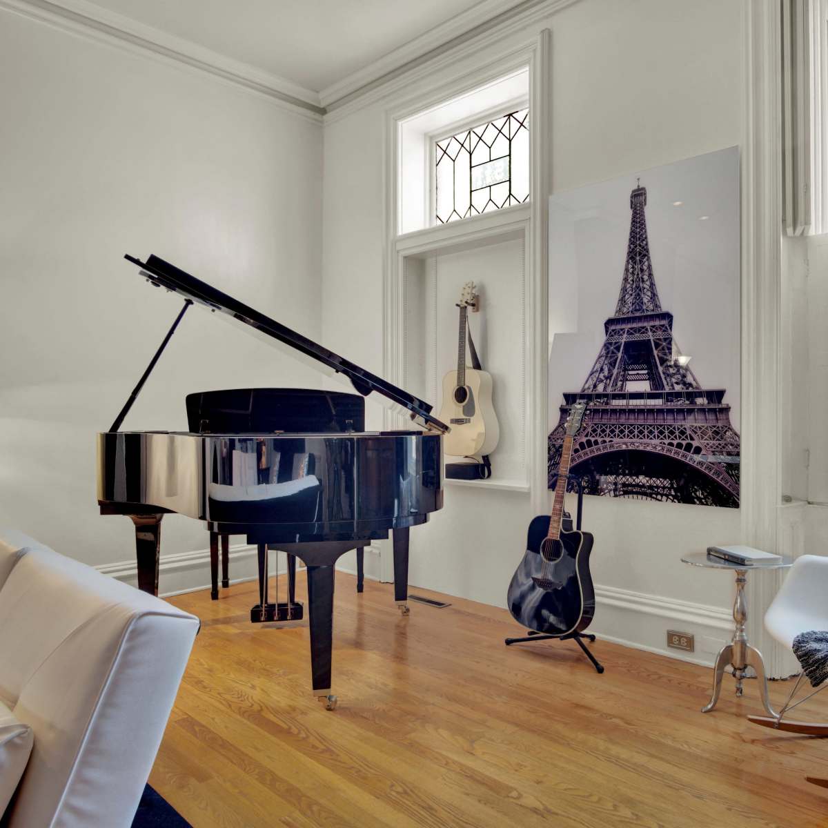 A grand piano sits beside a wall featuring a black-and-white photograph of the Eiffel Tower, with an acoustic guitar and a chair nearby in a well-lit room.