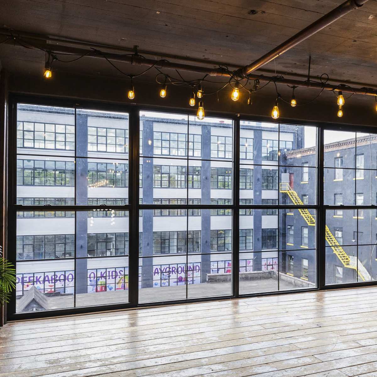 A spacious interior room features large windows overlooking a neighboring building with yellow fire escape stairs.