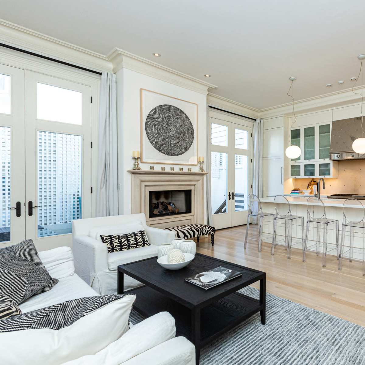 A bright, modern living room with a white sofa, black coffee table, and a kitchen featuring white cabinetry and a central island with bar stools.