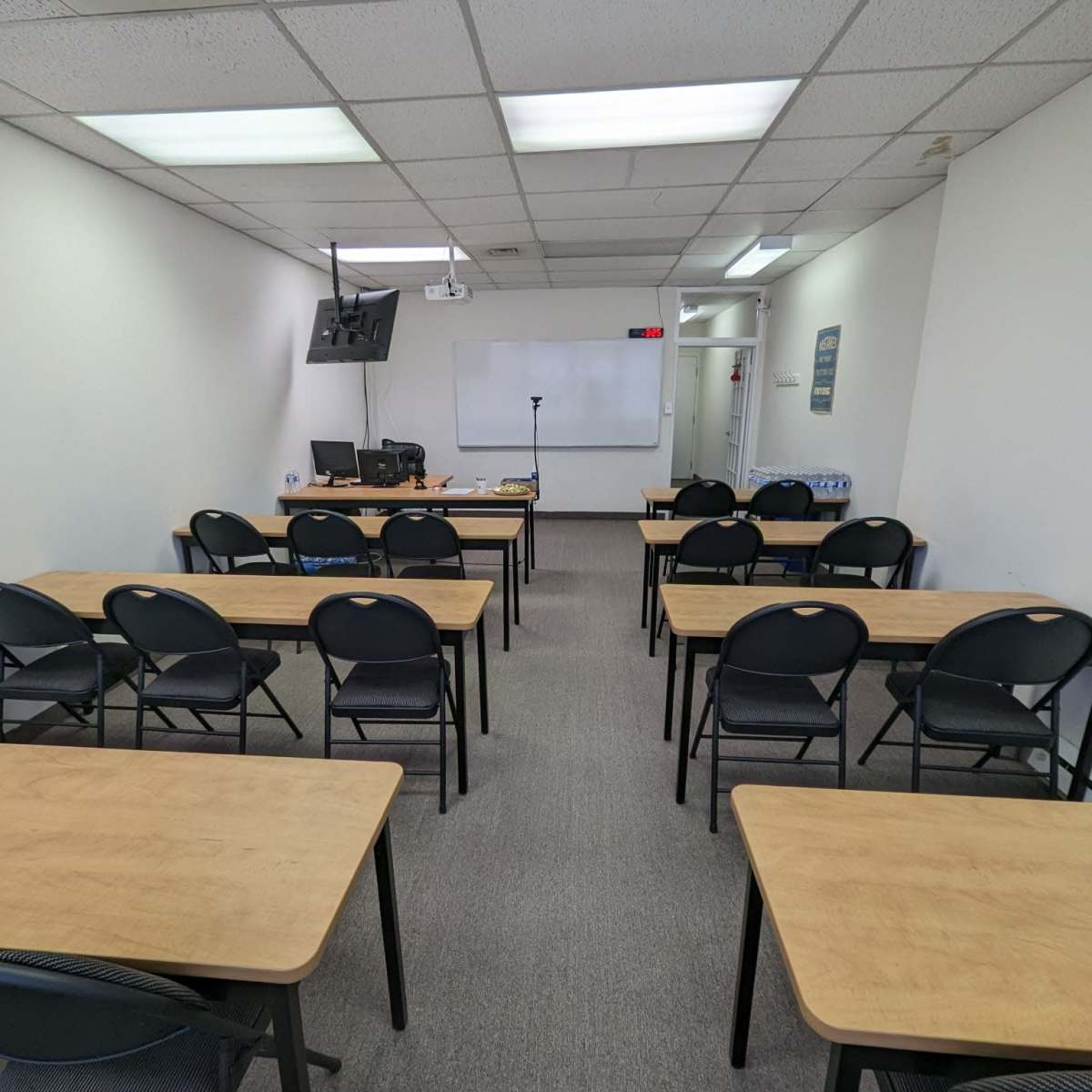 A classroom with rows of tables and chairs, a whiteboard, and a monitor at the front.
