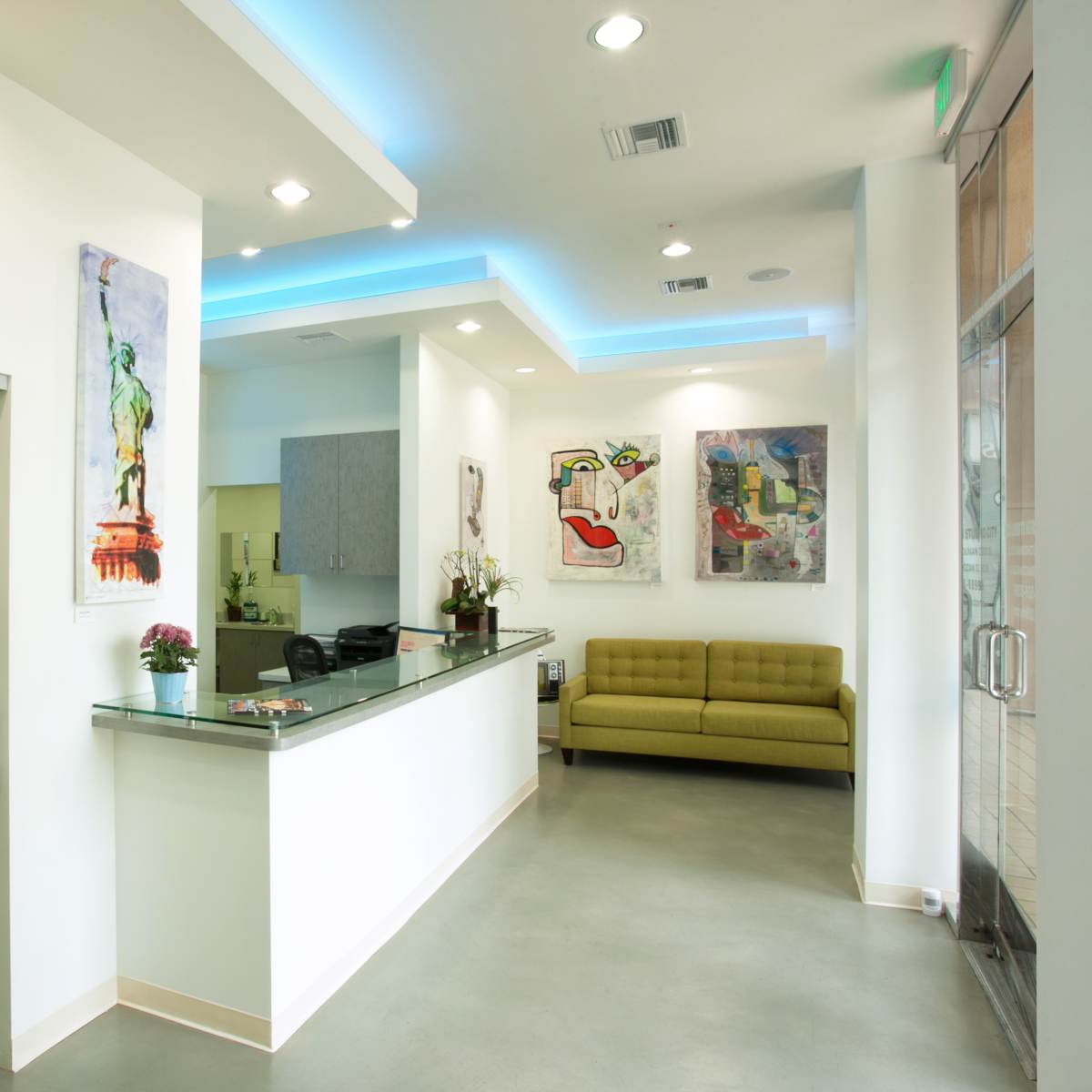A modern reception area with a glass door, a front desk, a green couch, and colorful artworks on the walls.