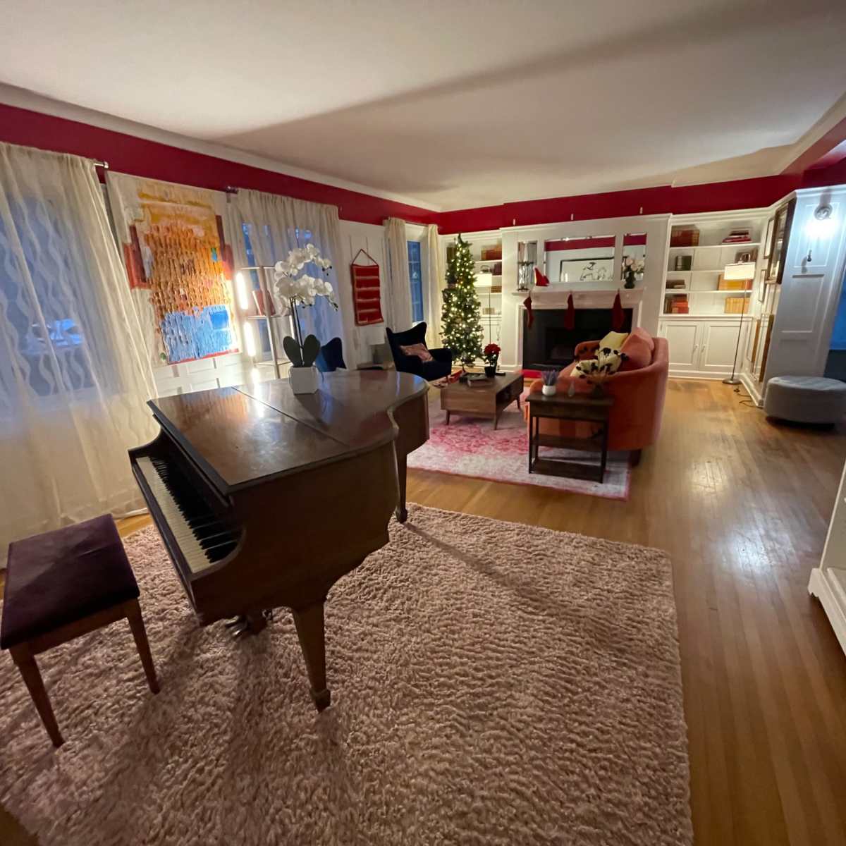 A spacious living room with a grand piano, a large area rug, and decorated shelves, featuring a Christmas tree and a cozy seating arrangement.