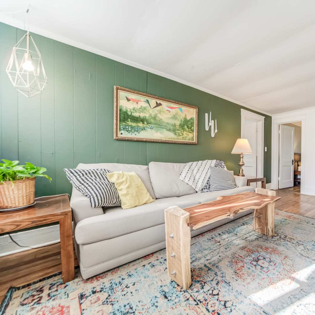 A living room featuring a light gray sofa with decorative pillows, a wooden coffee table, and a patterned rug, all arranged around a wall painted in green with artwork.