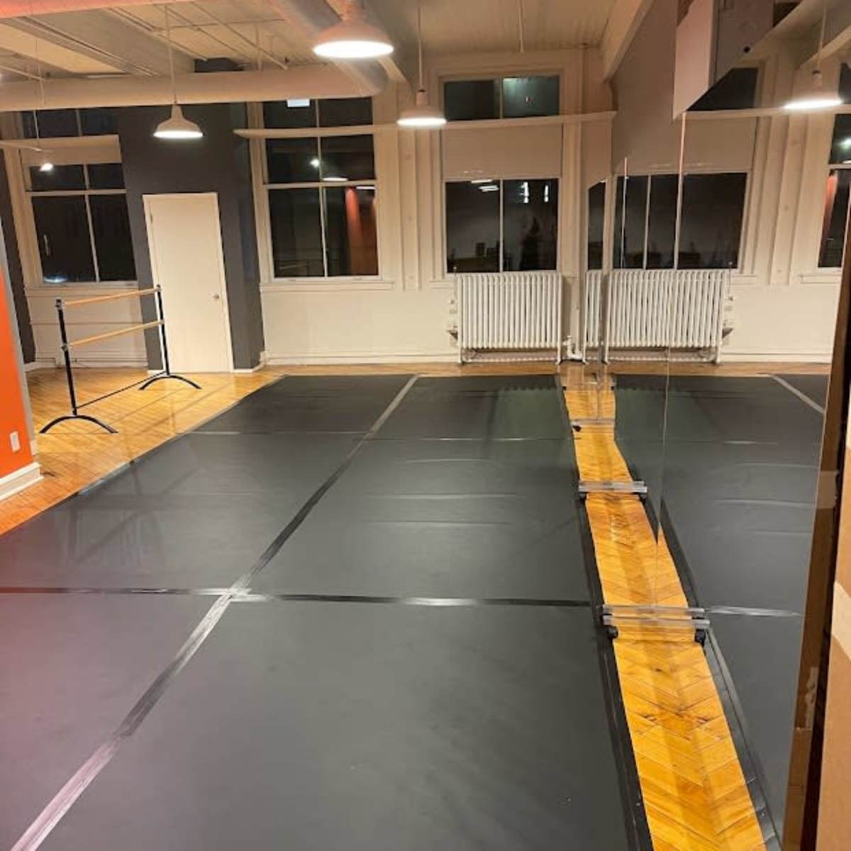 A dance studio with a wooden floor, black mats laid out, mirrors on one wall, and large windows allowing light in.