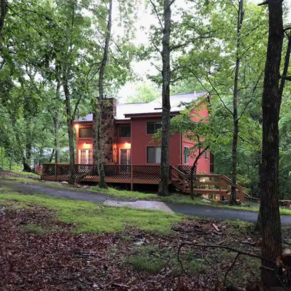 A two-story, pink house with a wooden deck sits among dense trees on a grassy lot.