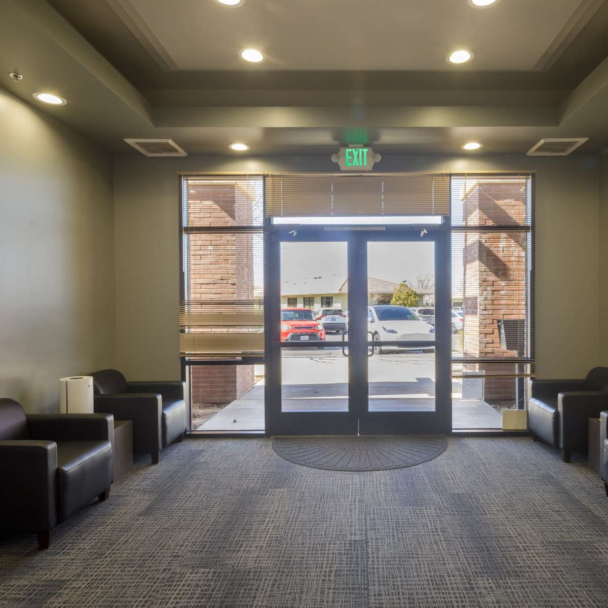 A modern waiting area with four black chairs arranged around a circular carpet, facing a set of double glass doors that lead outside.