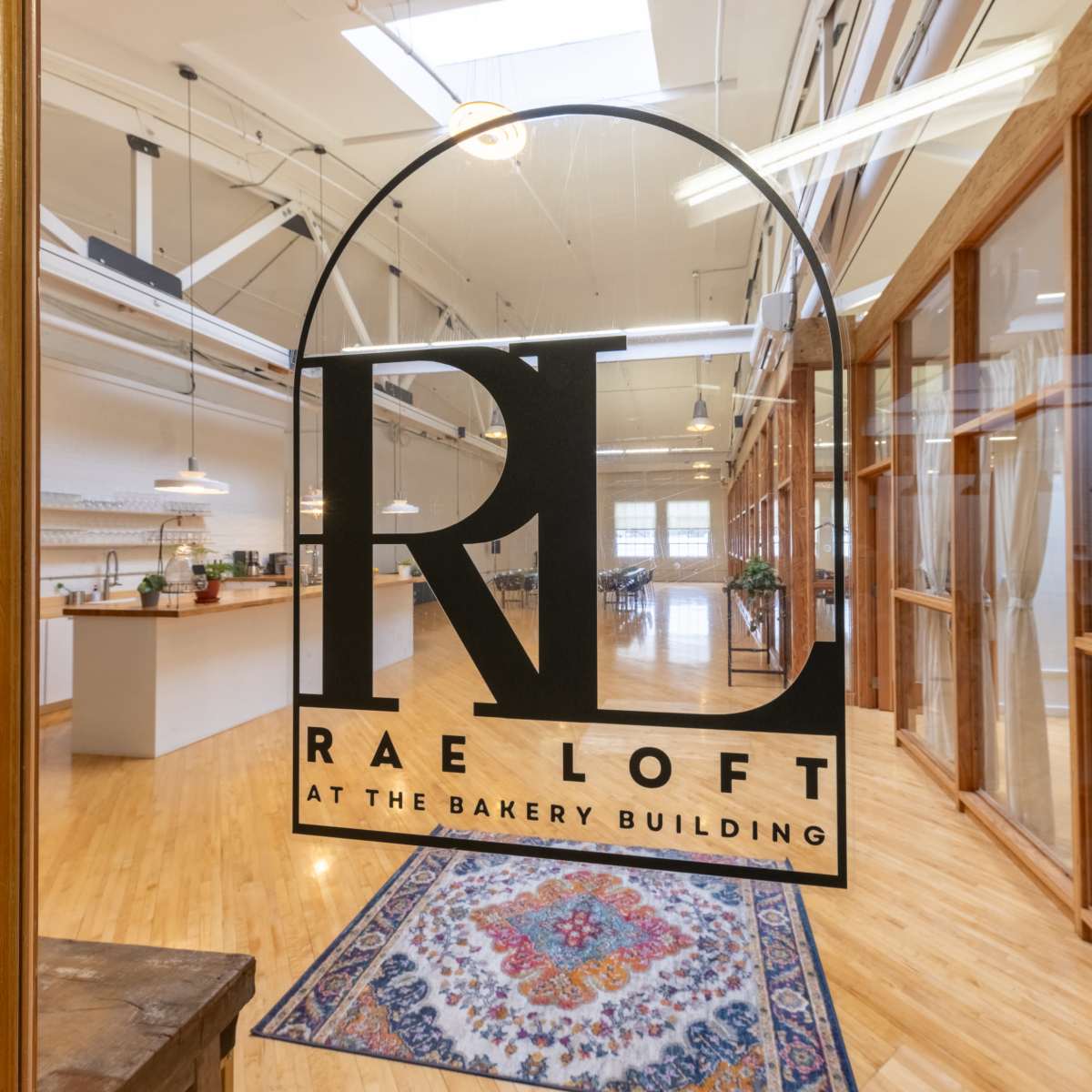 The entrance of a spacious room labeled "Rae Loft at the Bakery Building," featuring wooden floors and large windows, with a decorative rug in the foreground.