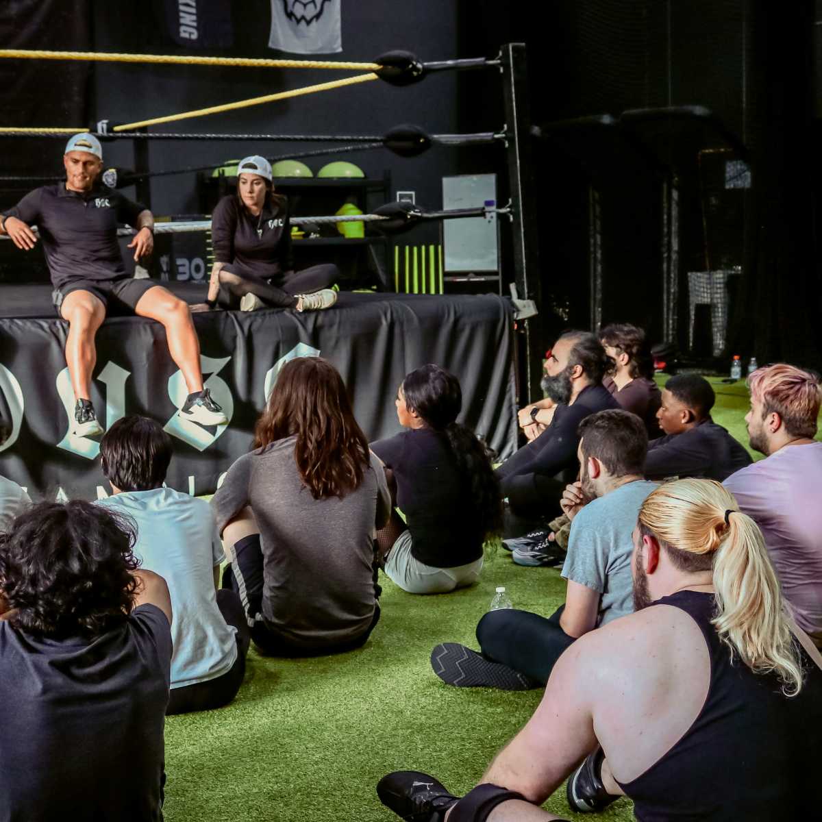 A group of individuals sits on a gym floor, attentively listening to three speakers seated on a wrestling ring.