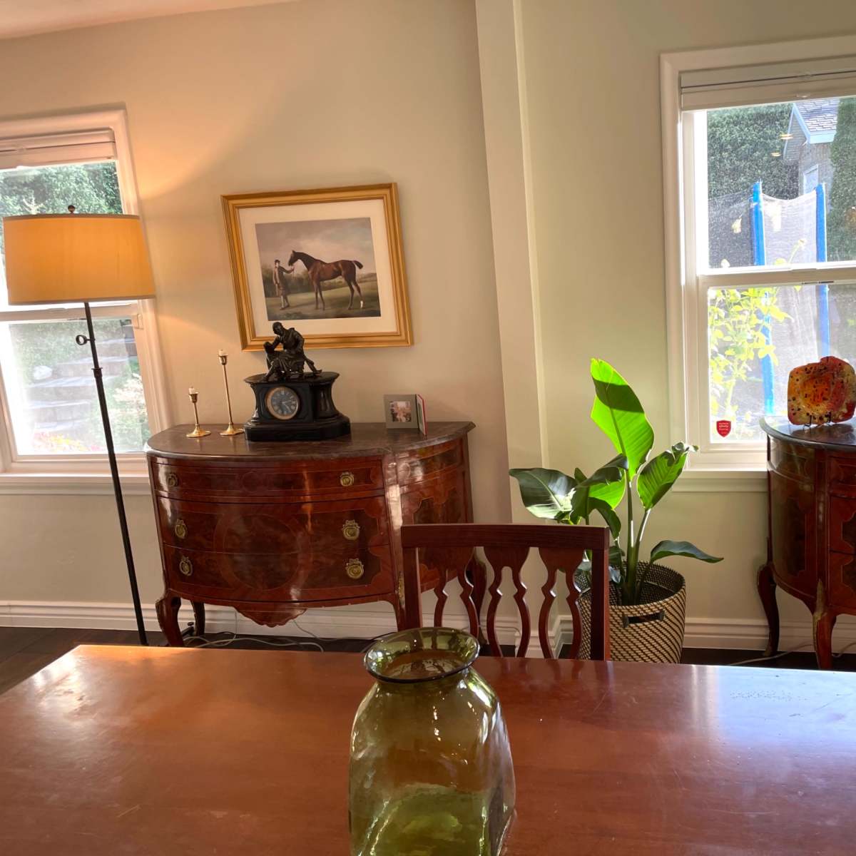 A well-lit room with two ornate side tables, a lamp, a painting of a horse, and a large plant beside a wooden dining table.