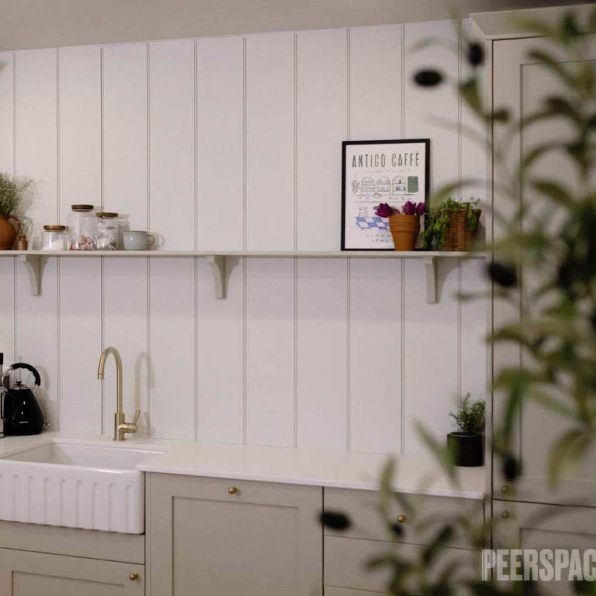 A modern kitchen with light-colored cabinetry, a farmhouse sink, and a shelf displaying plants and kitchenware against a paneled wall.