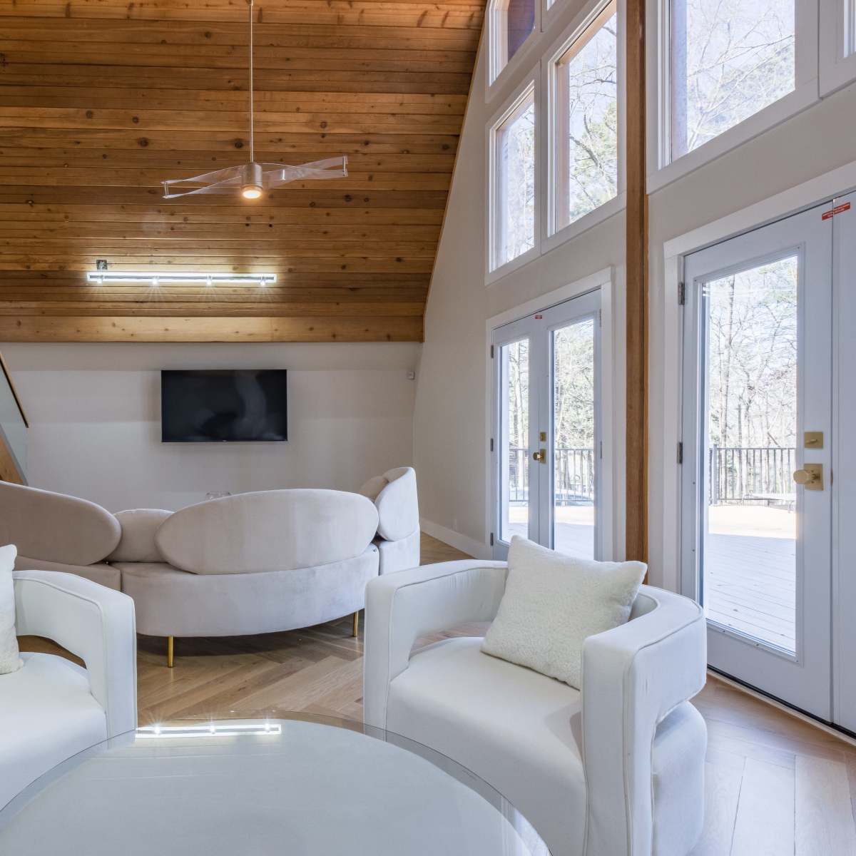 The interior features a modern living space with a wooden ceiling, a staircase, a circular sofa, and a glass table adjacent to large glass doors leading to an outdoor deck.