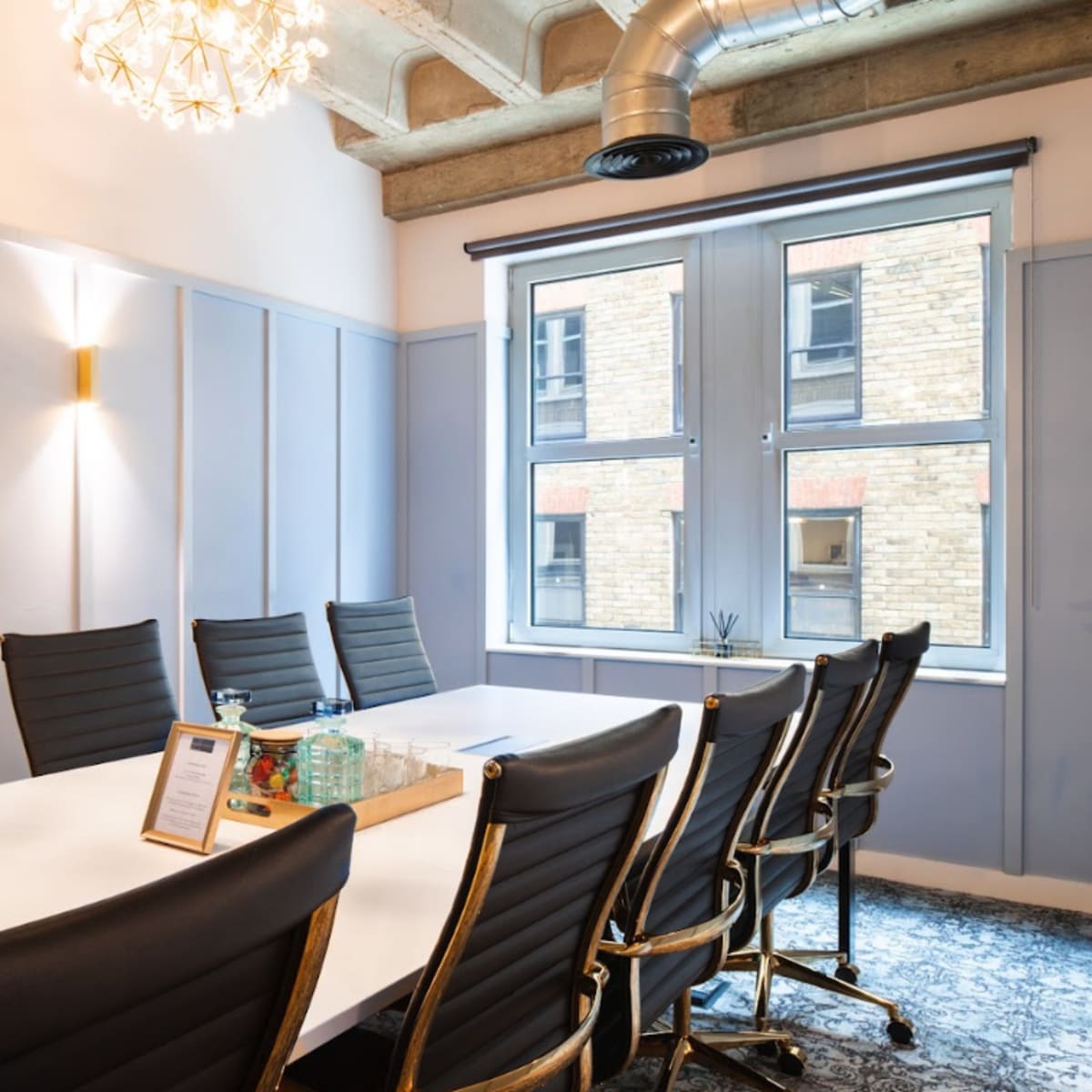 A conference room features a long white table surrounded by black leather chairs, illuminated by overhead lighting and natural light from large windows.