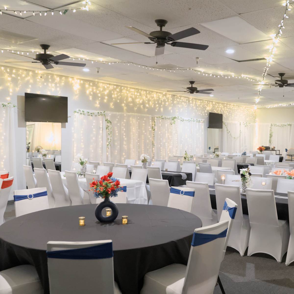 A banquet hall featuring rows of tables dressed in black and white linens, adorned with centerpieces, and illuminated by string lights overhead.