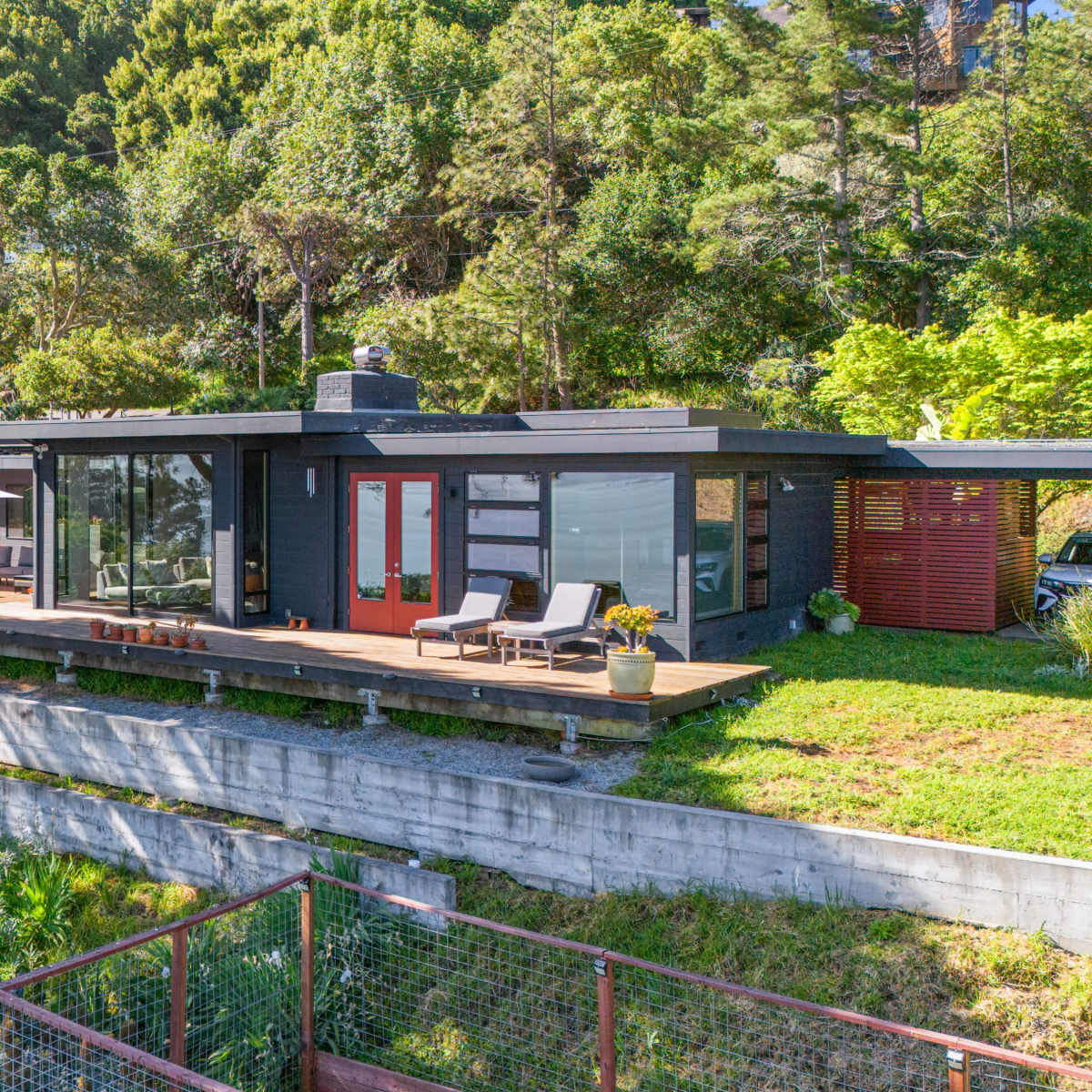 A modern house with large glass windows and a wooden deck, situated on a hillside surrounded by greenery.