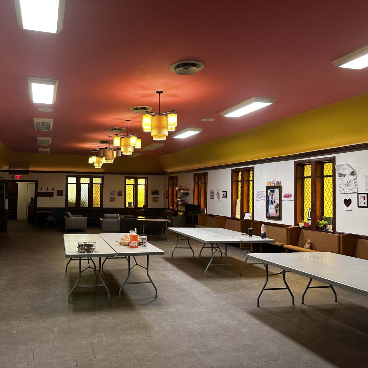 A spacious indoor room with tables arranged in the center, chairs along the walls, and decorative lighting above.