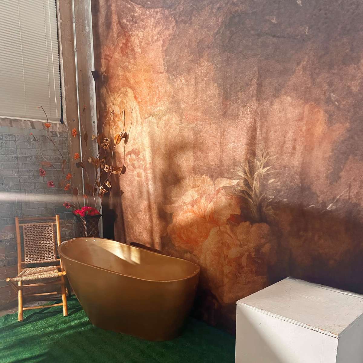A small bathroom setup with a golden tub, a white cube table, and a bamboo chair against a floral wall backdrop.