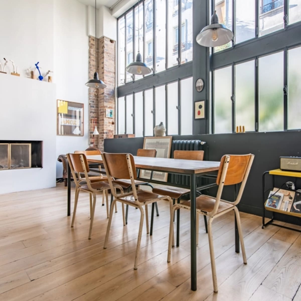 A dining area features a wooden table surrounded by chairs, large windows allowing natural light, and a fireplace against one wall.