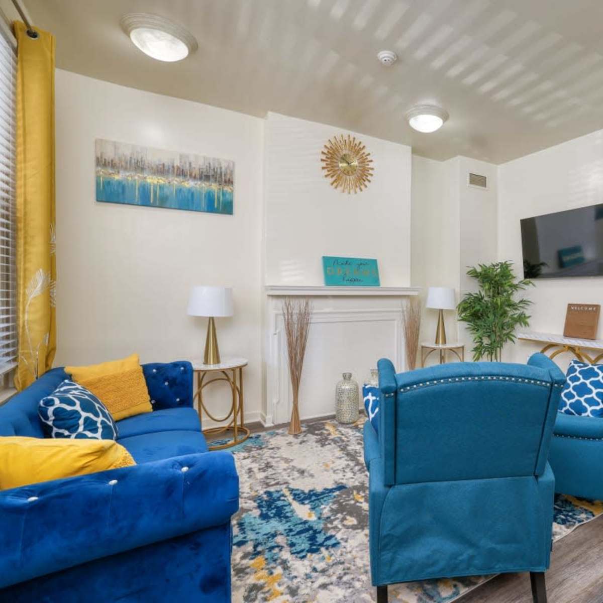 A living room with a blue sofa, two teal chairs, a patterned rug, a television on the wall, and a decorative fireplace.